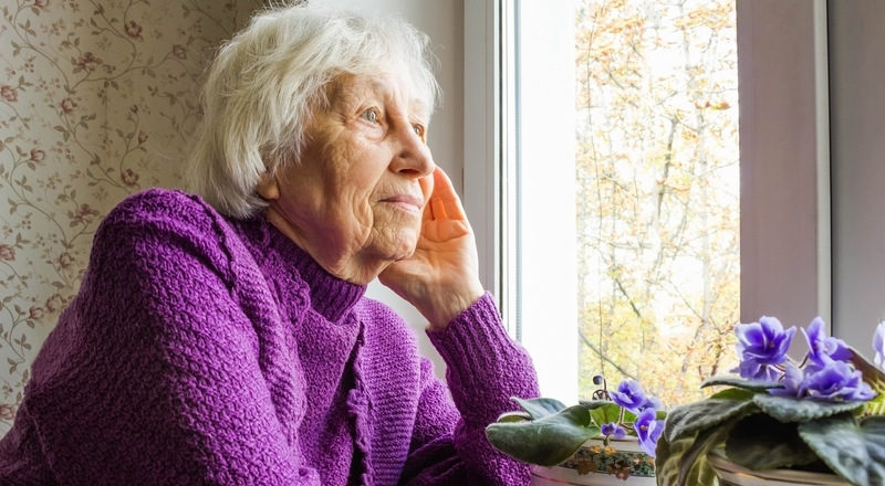 Äldre kvinna tittar ut genom fönster. Foto.