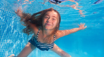 Flicka under vattnet i en simbassäng. Hon tittar in i kameran och ler. Foto.