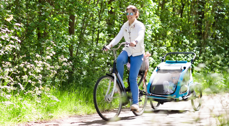 Kvinna cyklar en sommardag och har en cykelkärra med barn efter cykeln. Foto.