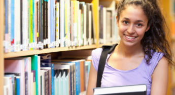 Mörkhårig ung kvinna står med böcker i famnen i bibliotek. Foto.