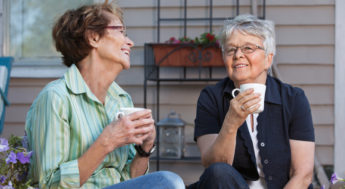 Två äldre, glada kvinnor samtalar och dricker kaffe på altan. Foto.
