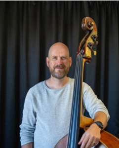 Man med skägg står med kontrabas. Foto.