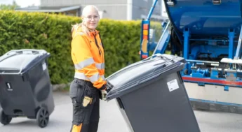 Kvinna i orange varseljacka som håller i en svart soptunna. Foto.