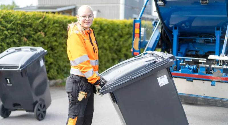Kvinna i orange varseljacka som håller i en svart soptunna. Foto.