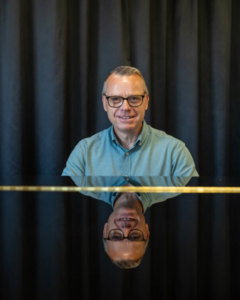 Man med glasögon sitter vid piano. Foto.