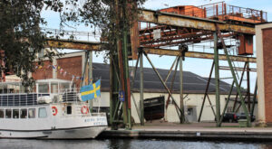 Kanalbåten Kung Sverker i kanalen med lyftkranar och byggnader i bakgrunden. Foto.