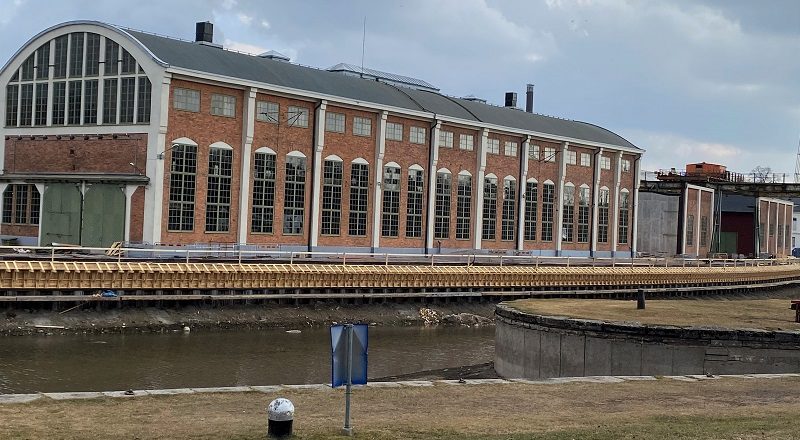 En gammal industrilokal i rött tegel med stora fönster, i förgrunden kajen till Göta kanal som renoveras.