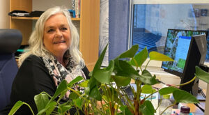 Kvinna sitter vid skrivbord. Gröna växter syns i förgrunden. Foto.