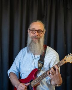 Man med skägg och glasögon håller i elgitarr. Foto.