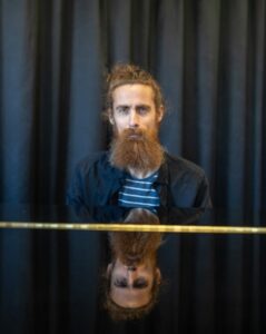 Man med skägg sitter vid piano. Foto.