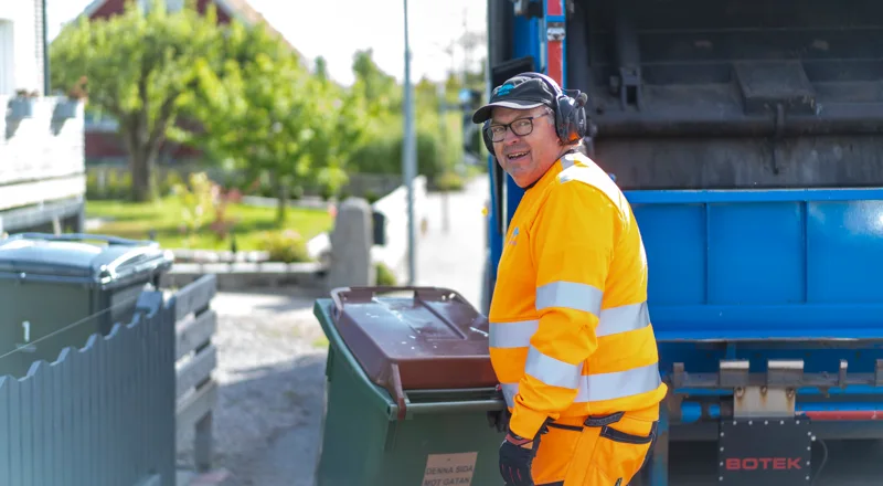 Man i orange varseljacka som står med en soptunna vid en sopbil. Foto.