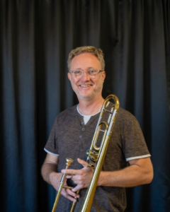 Man med glasögon håller i mässingsinstrument. Foto.