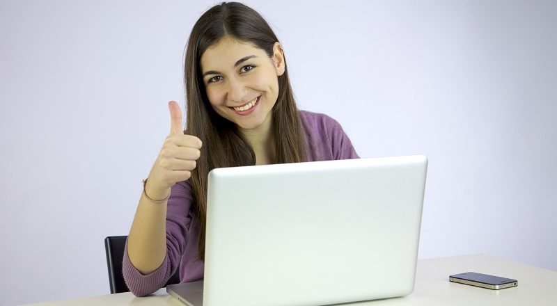 mörkhårig ung kvinna sitter vid laptop, ler brett och gör tummen upp