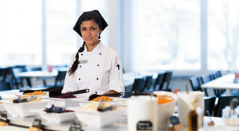 Kvinna med kockkläder står framför ett salladsbord. Foto.