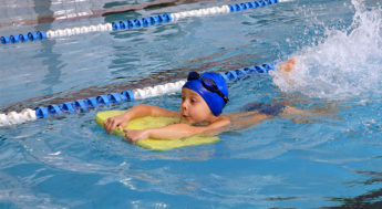 Bilden visar ett barn i en pool. Barnet simmar på mage och har hjälp av en gul flytplatta. Barnet har en blå badmössa och simglasögon på pannan.