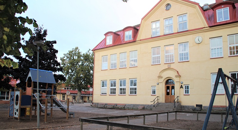 Stor gul stenbyggnad. I förgrunden syns lekredskap på skolgården.